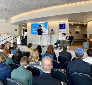 Three people on stage infront of a sitting audience. Photo.